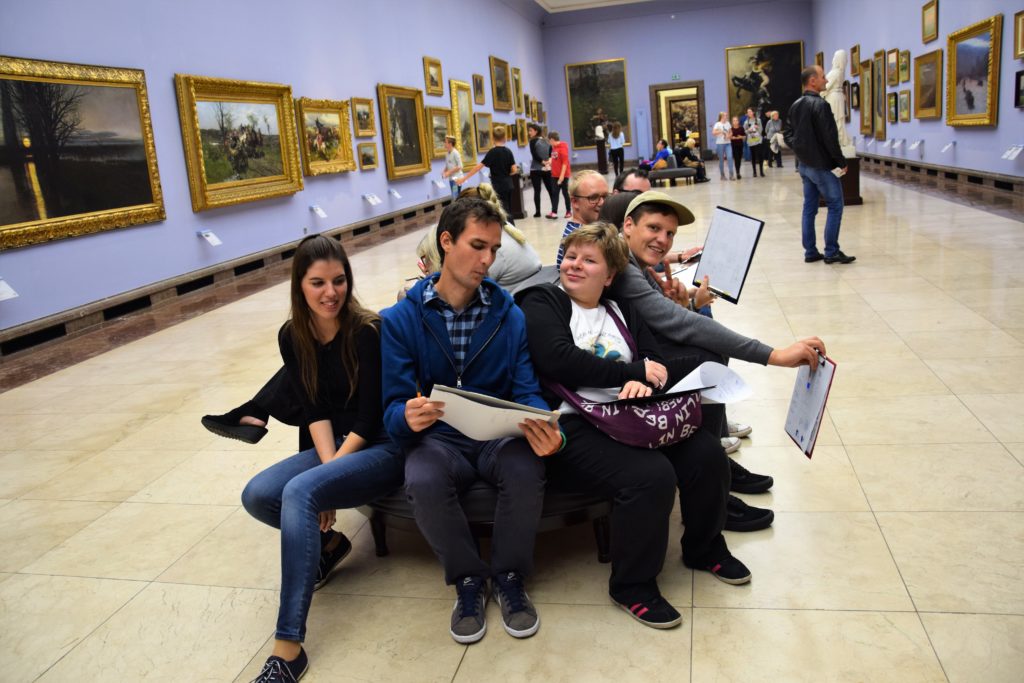 zdjęcie przedstawia grupę młodych osób w muzeum. Ludzie siedzą, pośród dzieł sztuki, w rękach trzymają kartki z tekstem – prawdopodobnie uczestniczą w lekcji muzealnej.