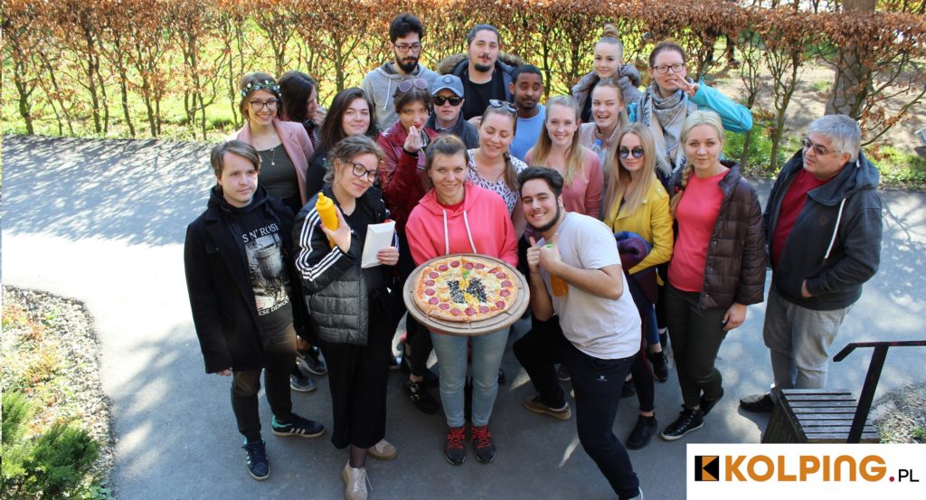 ZDJĘCIE 1 – zdjęcie przedstawia młodych ludzi w kolorowych strojach prezentujących upieczoną pizzę z logotypem KOLPING